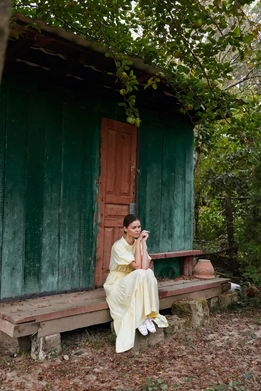 A woman in a pale yellow dress sits on a bench beside a green shed, surrounded by greenery and vines in a tranquil outdoor setting