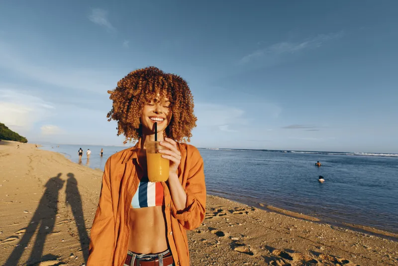 Woman, beach, sea, sunshine, smiling, curly hair, holding drink, summer, vacation, outdoors, warm jacket, bikini, golden sand, distant swimmers, blue sky, relaxed stance, natural colors