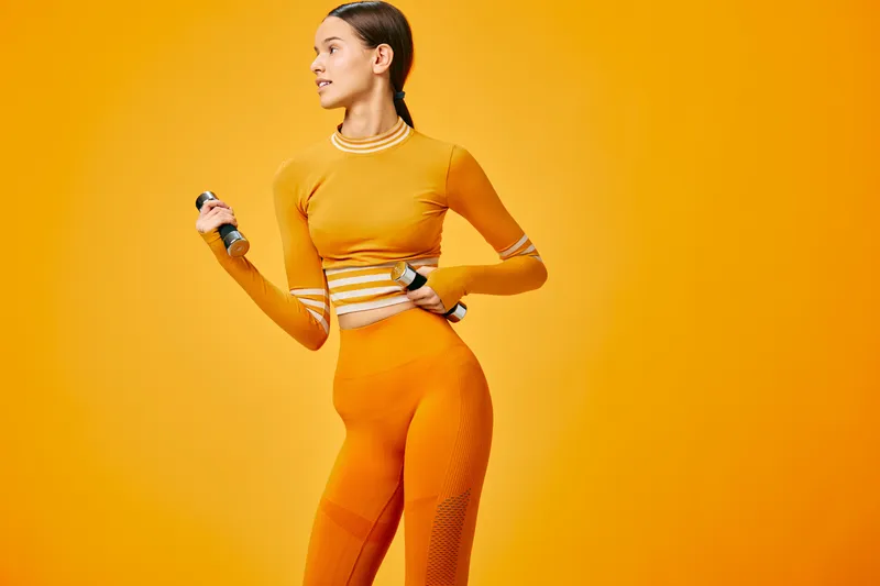 Woman in orange athletic wear holding a dumbbell, performing a dynamic workout in a vivid orange studio, showcasing strength, balance and energetic fitness.