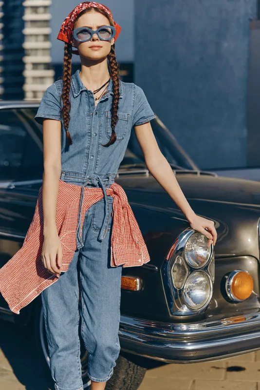 Lifestyle woman in modern boho-western denim street style with braided hair and sunglasses, wrapped in a checkered shirt, leaning on a vintage car under natural light.
