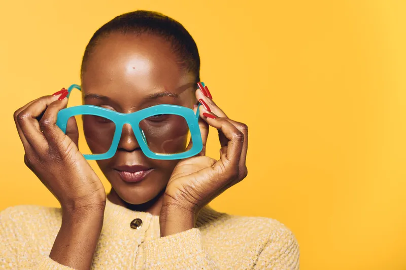 Close-up of a confident dark skinned African woman holding large blue oversized glasses on a yellow background. Concept of fashion, beauty, and bold accessories for women.