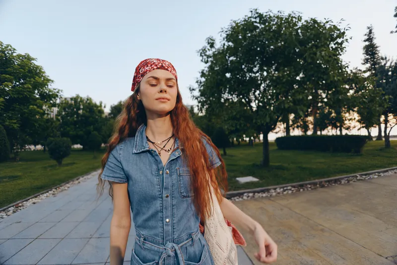 young woman with red bandana enjoys outdoor walk in casual denim outfit surrounded by green trees under clear sky on sunny day