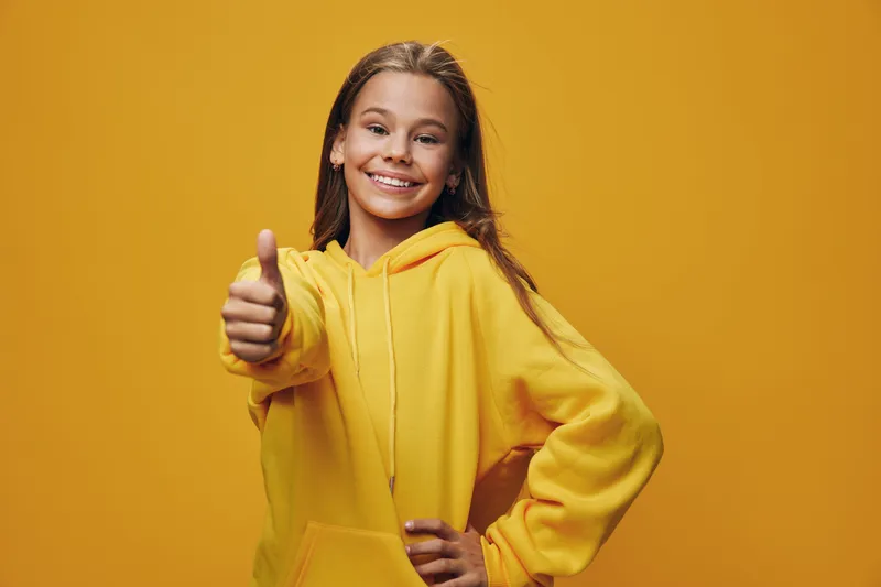 Girl in yellow hoodie smiling and showing thumb up gesture with confidence on yellow background. Positive expression, casual wear, and cheerful mood for success and encouragement.
