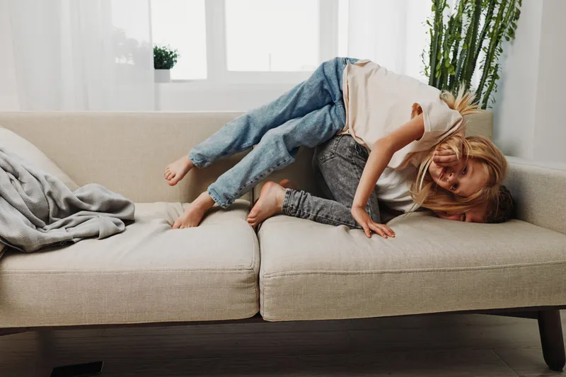 Happy mother and child playfully lying on beige couch in bright living room, enjoying family time and laughter together at home.