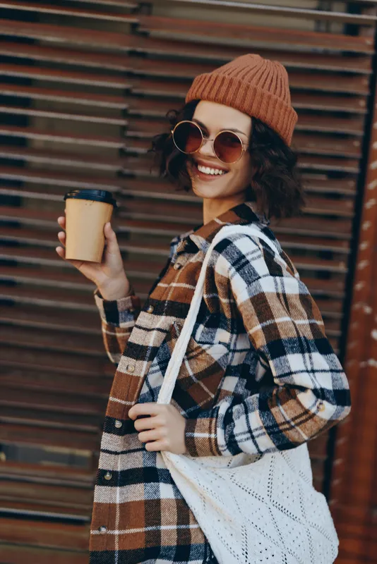 Woman holding coffee with a bright smile on a city street wearing plaid coat, beanie and sunglasses; candid fashion portrait with golden hour glow, authenticity, mindful living and emotional