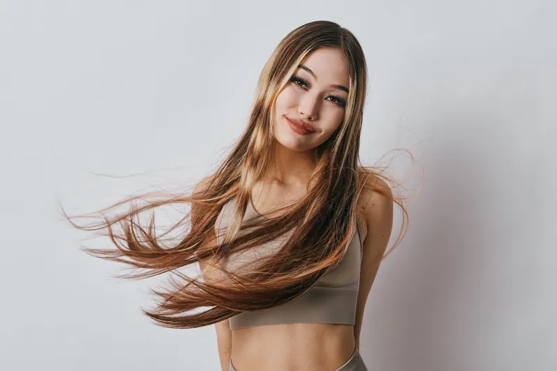 Woman with long flowing hair and sporty outfit smiling and looking at camera on white background. Female model with natural makeup and relaxed pose indoors.