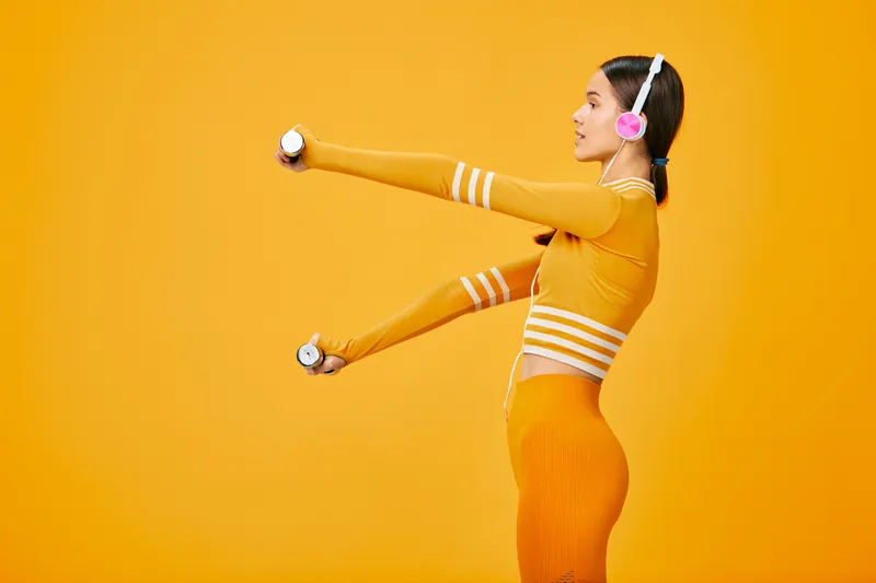 Woman in orange sportswear with headphones performs controlled arm raises using dumbbells against a vivid orange backdrop, capturing a dynamic fitness moment, strength and focused energy for an