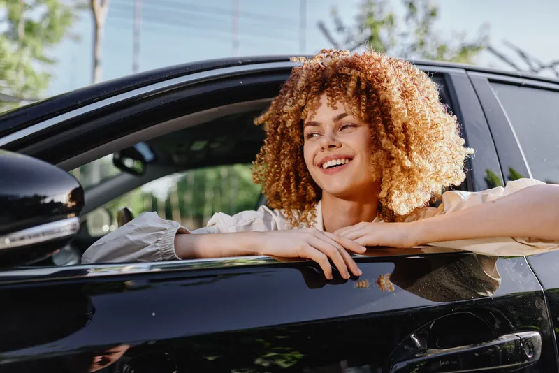 smiling woman sitting in black car with curly hair outdoors, enjoying sunny day and fresh air, casual style and happy mood conveying relaxation and freedom
