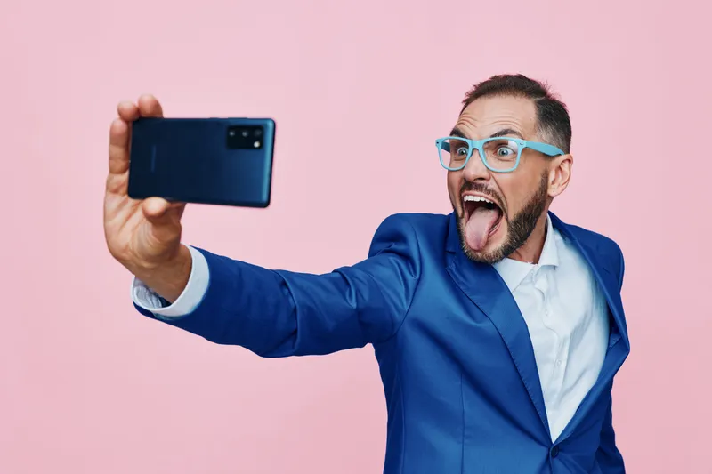 Professional man in a blue suit on a solid pink background. He exudes confidence and energy while taking a selfie, showcasing modern work life and dynamic emotion in a clean, isolated color setting.