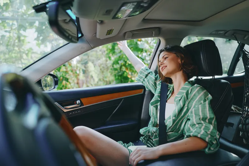Young woman driver sits in the front seat of a car, smiling and relaxed, wearing a green striped shirt as daylight streams through the window.
