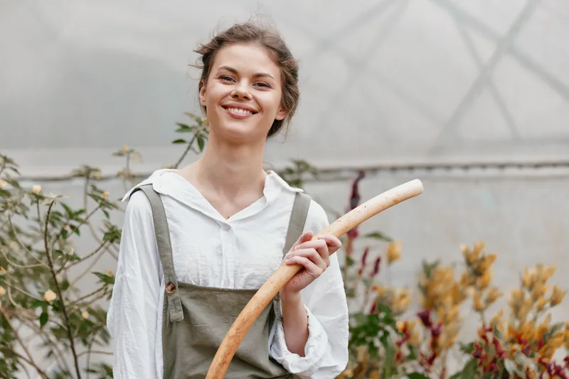 Happy young woman in a green garden holding a wooden stick, wearing casual grey overalls and a white shirt, emphasizing a cheerful, eco friendly lifestyle