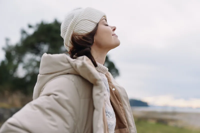 Woman, portrait, outdoor, winter, coat, beanie wearing and looking up toward the sky with a calm expression in a natural coastal landscape.