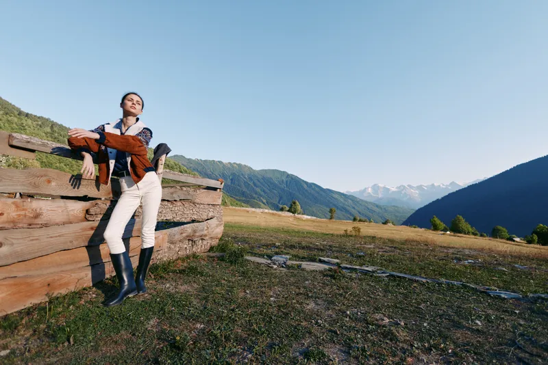 Woman leaning on wooden fence in sunny mountain meadow, wearing riding boots and holding jacket, relaxed countryside portrait with expansive alpine view and clear blue sky.