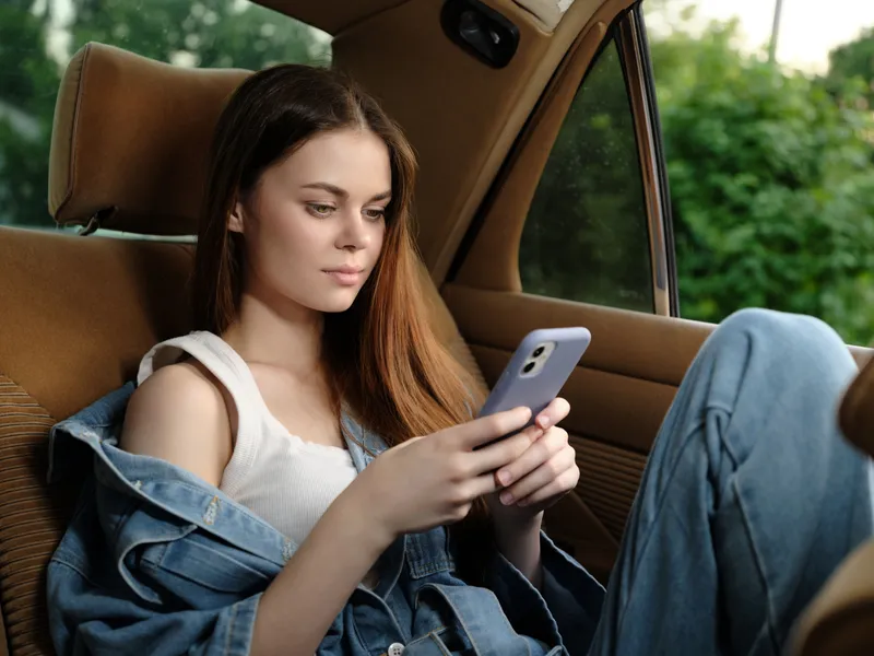 Young woman in casual denim outfit using smartphone in a car, expressing focus and relaxation, surrounded by greenery in soft daylight.