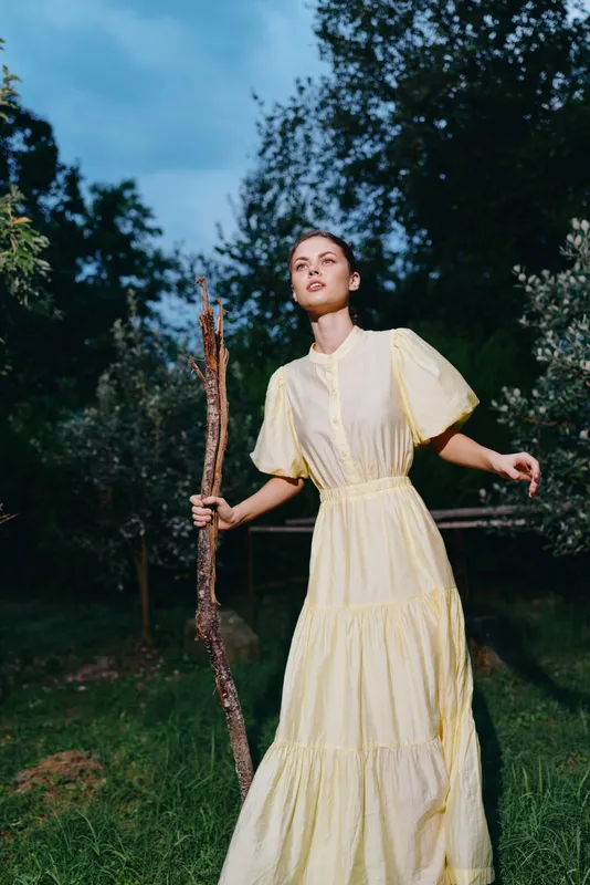 Woman in a pale yellow dress stands in a forest clearing at dusk, holding a wooden staff, exuding calm, ethereal atmosphere and a connection to nature