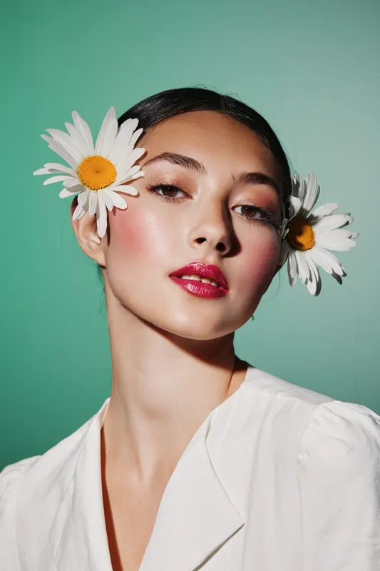 Portrait of a fashionable young woman with fresh daisies in her hair against greenale background. Glossy lips and soft glowing skin create floral beauty concept.