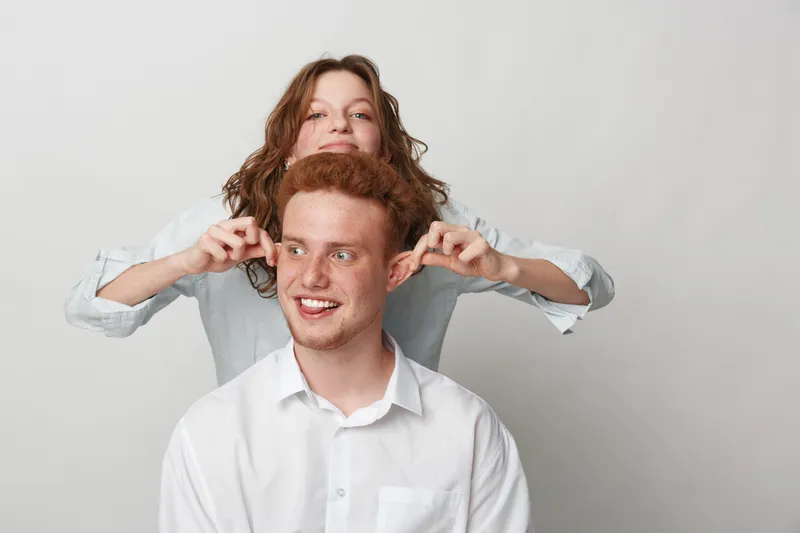 Playful young woman pulls ears of smiling redhead man in white shirt. Studio portrait on light background. Concept of friendship, fun, humor, and casual lifestyle.