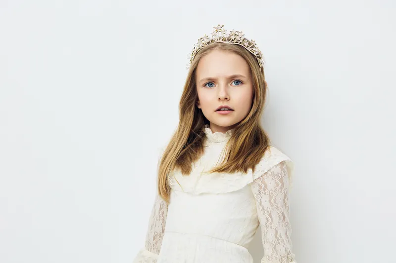 Young girl in a white lace dress and tiara, exuding elegance with a serene expression against a light background, perfect for children s fashion