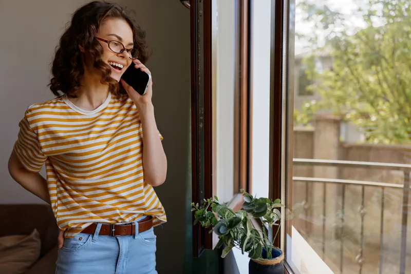 A young woman with curly hair stands by a sunlit window in a striped t shirt, talking on a mobile phone in a cozy home, exuding relaxed confidence.