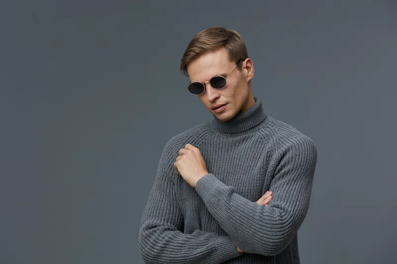 Stylish young man wearing gray turtleneck sweater and black sunglasses posing against plain gray background in a modern studio shot. Fashionable menswear, confident and trendy look, casual clothing, m