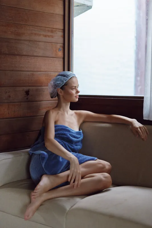Relaxed woman wrapped in blue towel sitting by window inside wooden room, natural light highlighting calm expression and serene atmosphere in cozy interior setting.