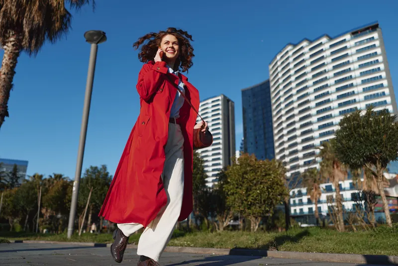 A woman in a bright red trench walks confidently along a sunny urban street, her coat fluttering as she strides past modern high rises and palm trees.