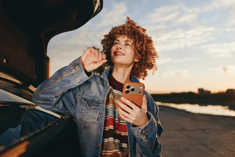Woman lifestyle outdoors by car at sunset, curly hair, denim jacket, holding phone and smiling, relaxed posture, warm golden light, soft scarf, sea horizon, light breeze