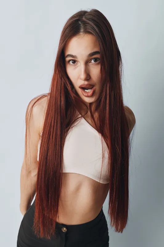 Young woman with long straight hair and surprised expression wearing a sleeveless top and black pants standing against white background in studio portrait.