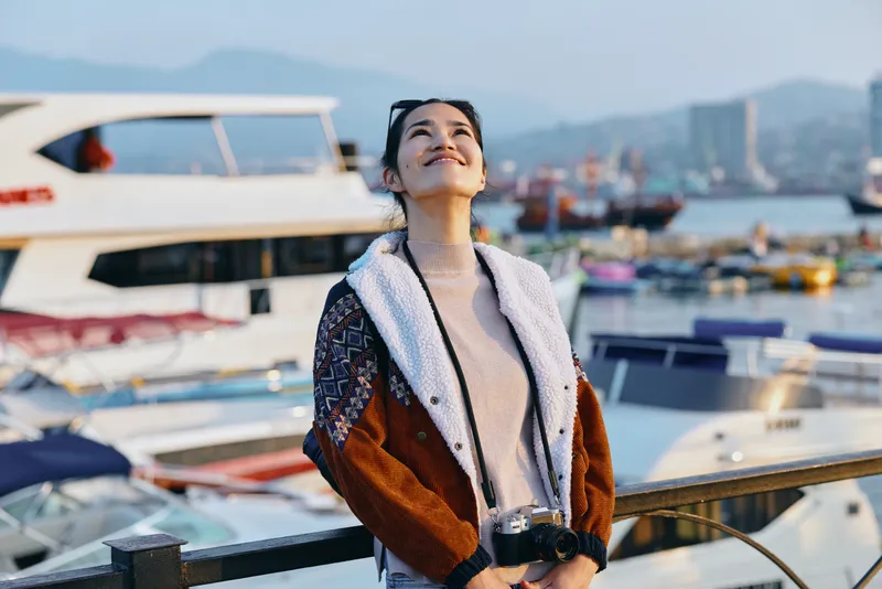Woman by a marina harbor in a stylish jacket, outdoor light, looking up with a relaxed smile toward boats and water, capturing travel style and seaside charm