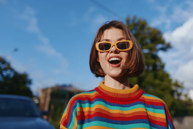 Young woman smiling with sunglasses and short hair in a colorful striped sweater, standing outdoors on a sunny suburban street, joyful expression, casual fashion and lifestyle travel vibe.