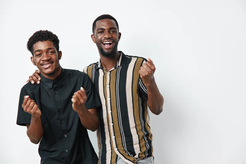Two young black men with dark skin smiling and celebrating together on white background. Friends, happiness, casual clothing, joy and positive emotions in studio.