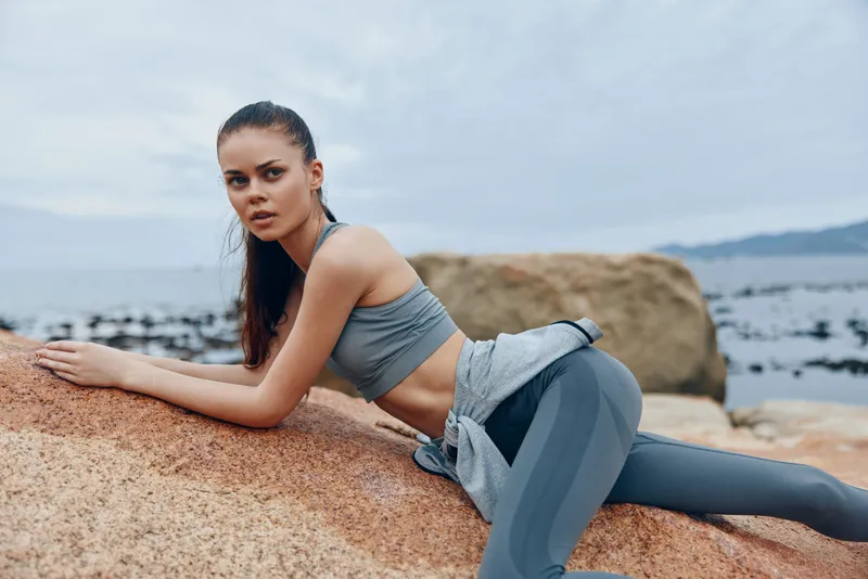 Young athletic woman in activewear poses on rocky beach, showcasing fitness and determination, against a serene ocean background in shades of blue and gray