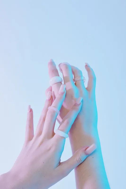 Close-up of female hands with long nails and multiple white rings on fingers. Soft blue lighting highlights beauty and fashion concept on a plain background.