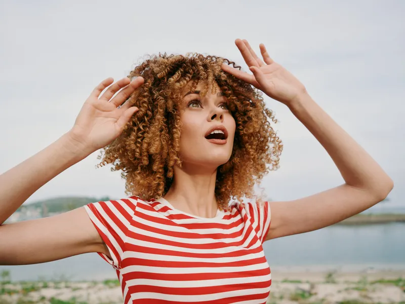 Joyful young woman with curly hair raises her arms beside a sunny beach, wearing a red striped shirt, capturing a carefree summer moment and an expressive, hopeful gaze.