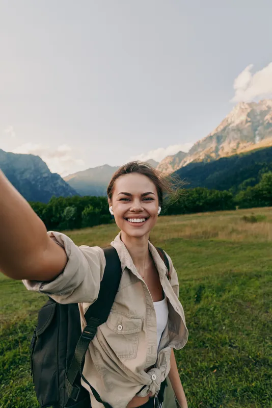 Young woman with a backpack takes a sunny outdoor selfie in a wide mountain meadow, smiling warmly as the scenery unfolds with a relaxed, adventurous vibe.