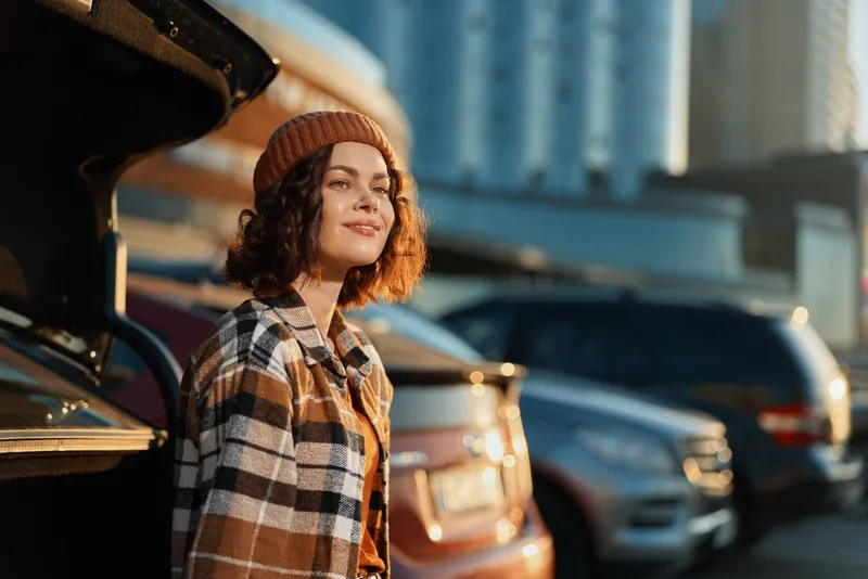 Woman by car in parking city, smile near open trunk in warm sunlight at golden hour glow. Authenticity and candid lifestyle scene of mindful living with emotional storytelling and urban vibe.