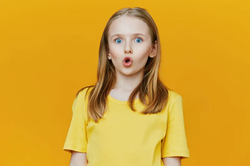 A surprised young girl with long blonde hair wearing a bright yellow shirt, set against a vibrant yellow background, expressing surprise and joy