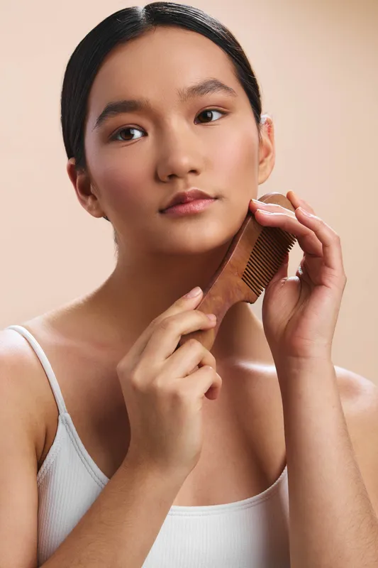 asian woman is seen grooming her hair with a wooden brush in a minimalist studio, calm expression, soft lighting, clean aesthetic, preparing for a daily beauty routine.