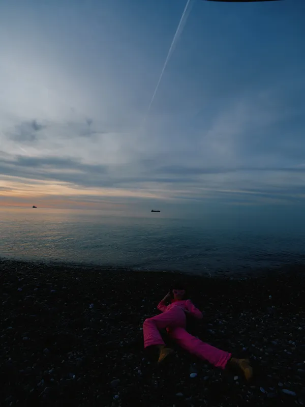 Person lying on a pebbled beach at dusk, pink clothing, calm ocean and distant boats, dramatic sky with a jet trail, serene outdoor scene for travel or nature photography.