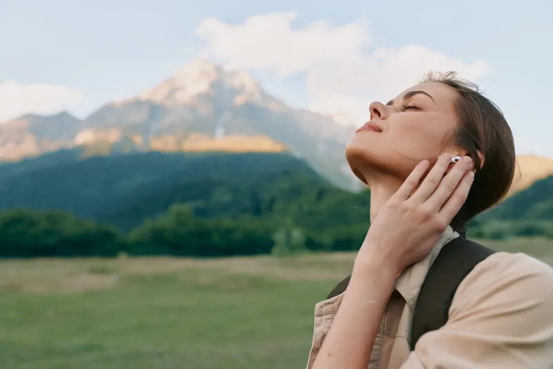 Young woman with long brown hair enjoys fresh mountain air with closed eyes and relaxed expression in a green field under a clear sky during daytime.
