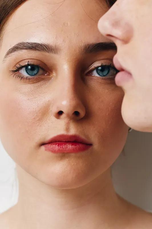 Portrait of woman with blue eyes and red lips looking at camera while man profile is near her face. Closeup beauty skin with natural makeup and facial expression emotion in studio light.