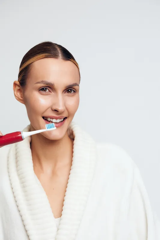 Smiling woman in white bathrobe brushing her teeth with a red toothbrush on a light background. Concept of dental hygiene, morning routine, and personal care with natural lighting.