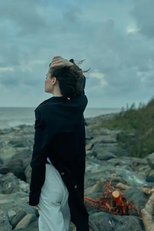 Woman in a windy seascape on a rocky coastline, coat fluttering in the breeze, gazing toward the ocean under a moody cloudy sky, conveying outdoor fashion and solitude, a dramatic coastal moment