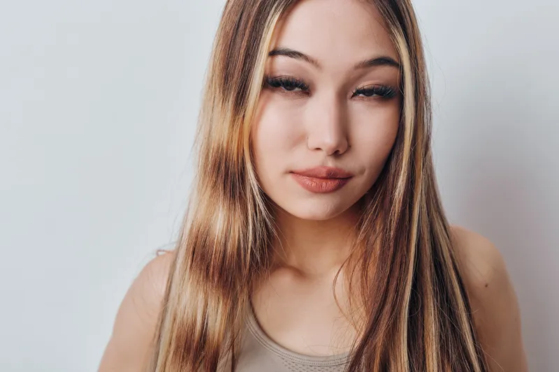 Young woman with long hair wearing a sleeveless top posing confidently in front of a light background. Portrait, beauty, natural look and casual style with calm expression.