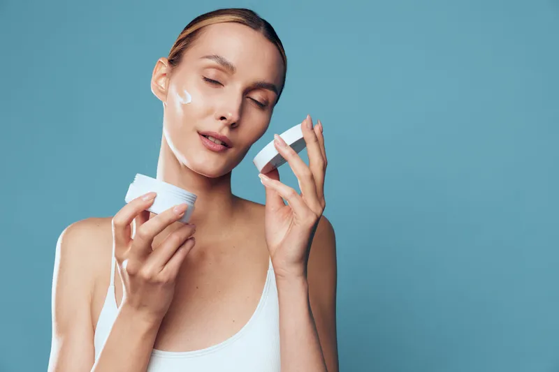 A young woman applies moisturizer from a cream jar onto her face, standing against a soft blue background, capturing a calm skincare moment with focused, natural expression.