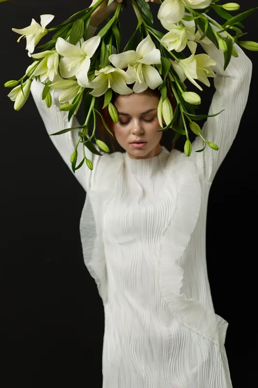 A serene woman in a flowing white dress gracefully holds a crown of white flowers, symbolizing purity and elegance in a minimalist setting