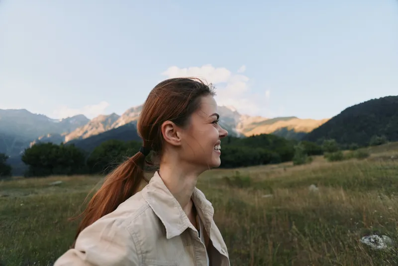 Young woman with long brown hair smiles while enjoying a scenic mountain landscape during a clear day in casual beige clothing.