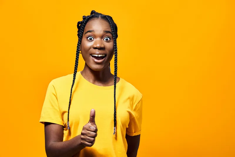 Joyful young African woman in a bright yellow t shirt giving a thumbs up against a vibrant orange background, expressing happiness and positivity