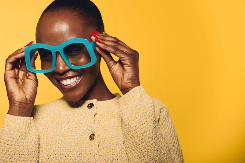 Happy African woman with dark skin wearing oversized blue glasses and beige knitted sweater smiles against bright yellow background. Concept of joy, fashion, and vibrant portrait.