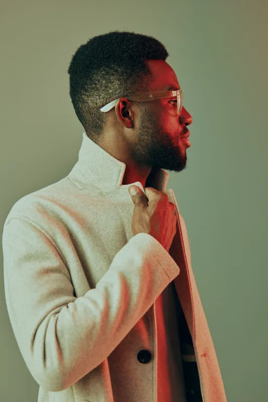 Stylish African man in a beige coat with glasses, expressing confidence against a minimalistic background, perfect for fashion and lifestyle themes